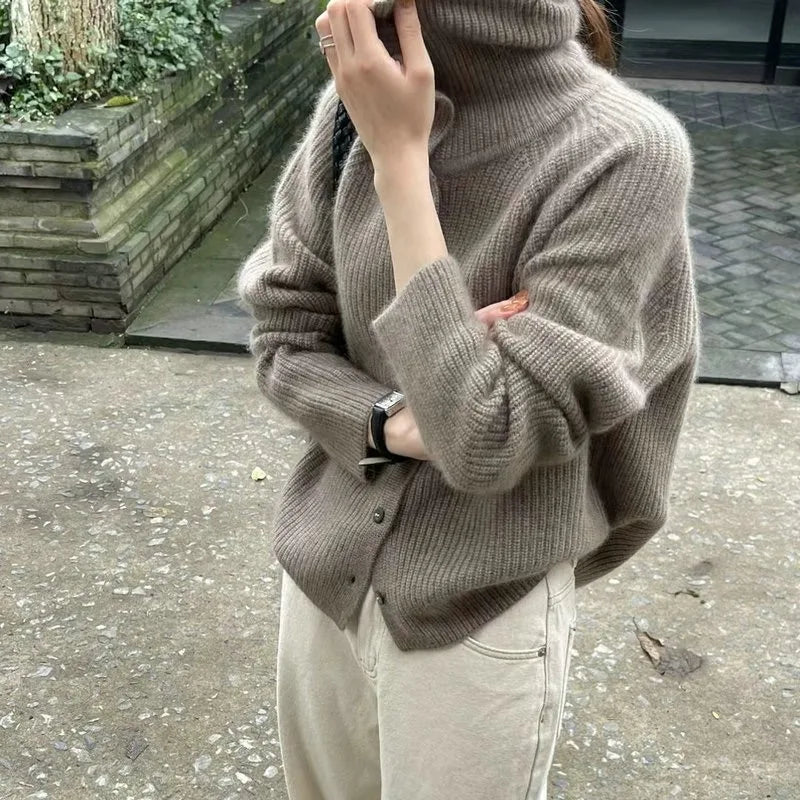 Femme portant un pull en laine beige à col roulé, manches longues, avec un pantalon beige, posant à l'extérieur près d'un mur en briques.