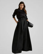 Femme portant une robe noire élégante avec manches longues et jupe ample, tenant une pochette. Mode chic, style minimaliste, tenue de soirée.