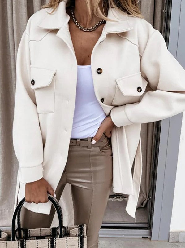 Femme portant une surchemise beige élégante avec boutons noirs, associée à un top blanc et un pantalon beige, tenant un sac à main rayé.