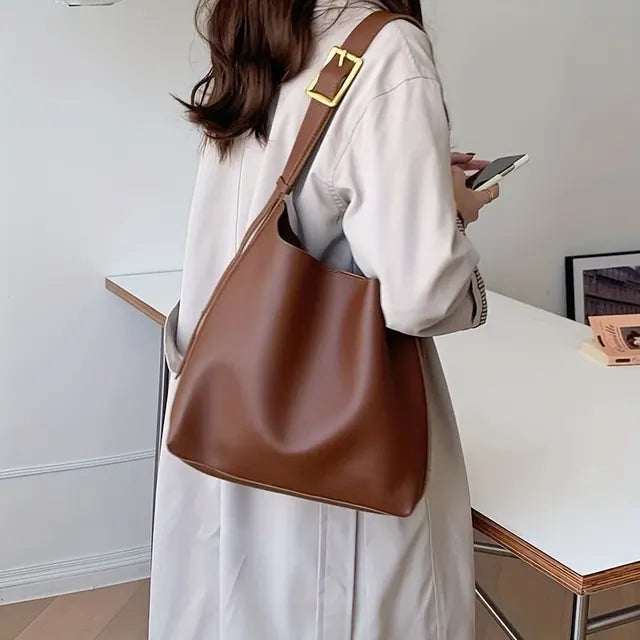 Femme en manteau beige avec sac à main en cuir marron, tenant un smartphone. Mode élégante, accessoires tendance, style automne-hiver.