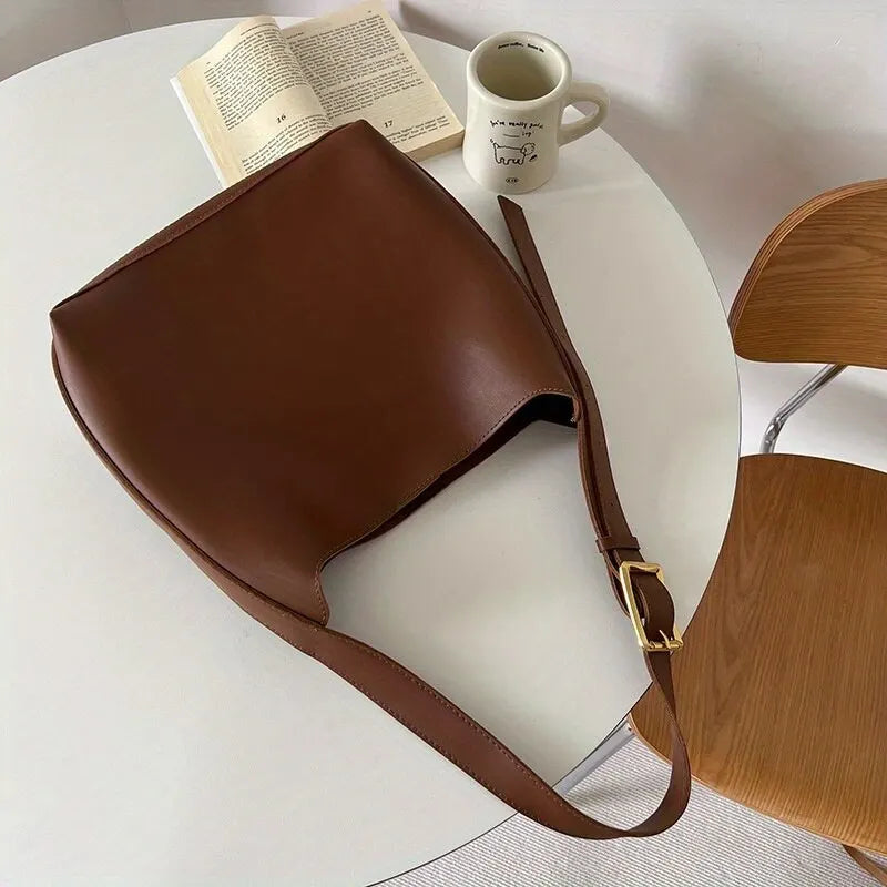 Sac à main en cuir marron sur une table blanche avec livre ouvert et tasse de café. Design élégant, accessoire mode tendance, style minimaliste.