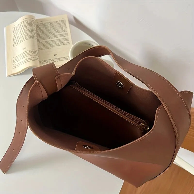 Sac à main en cuir marron ouvert sur une table blanche, avec un livre ouvert et une tasse de café à côté. Accessoire mode élégant et pratique.