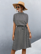 Femme en robe noire et blanche à motifs géométriques, chapeau de paille, lunettes de soleil, posant devant un mur gris. Mode estivale élégante.