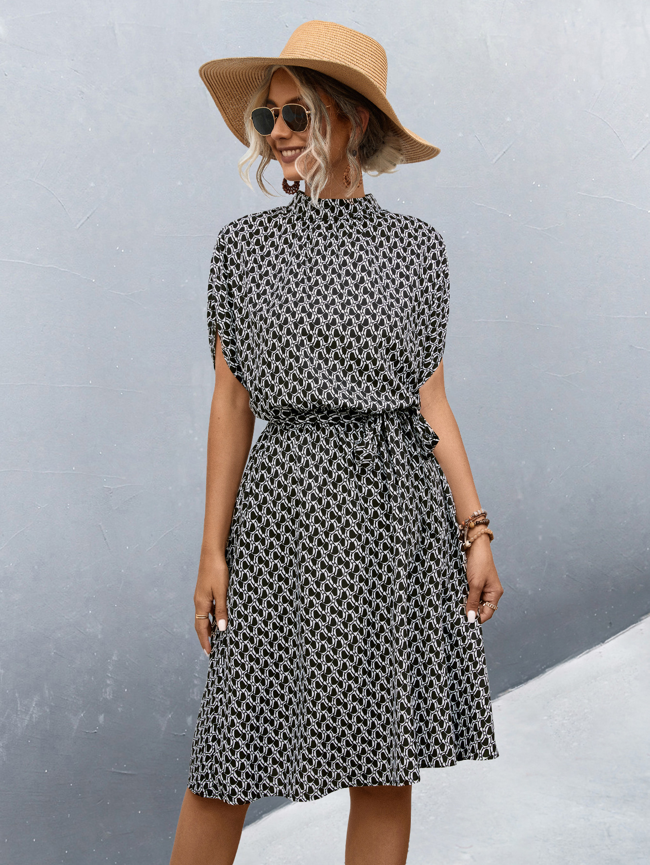 Femme en robe noire et blanche à motifs géométriques, chapeau de paille, lunettes de soleil, posant devant un mur gris. Mode estivale élégante.