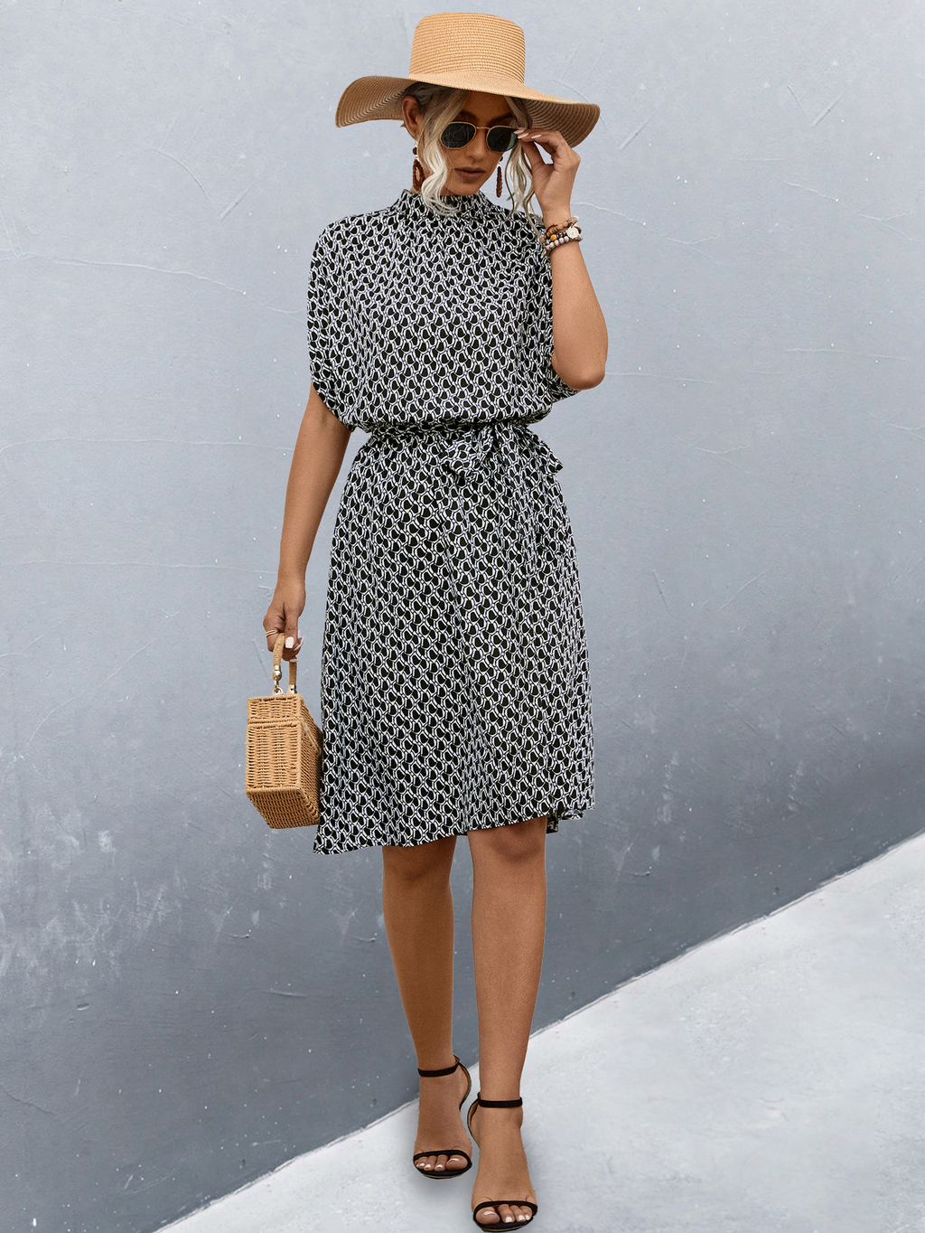 Femme élégante en robe à motifs noir et blanc, chapeau de paille, lunettes de soleil, sandales noires, tenant un sac en osier, posant contre un mur gris.