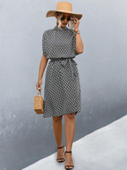 Femme élégante en robe à motifs noir et blanc, chapeau de paille, lunettes de soleil, sandales noires, tenant un sac en osier, posant contre un mur gris.