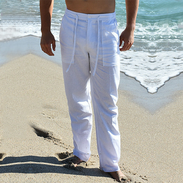 Homme en pantalon blanc en lin sur une plage, pieds nus sur le sable, avec vagues en arrière-plan. Mode estivale, tenue décontractée bord de mer.