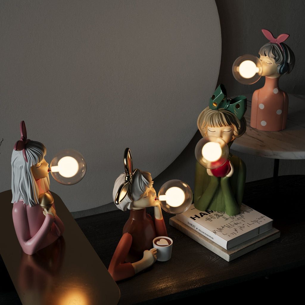 Quatre lampes décoratives en forme de personnages soufflant des bulles lumineuses, posées sur une table avec des livres, créant une ambiance artistique et moderne.