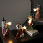 Quatre lampes décoratives en forme de personnages soufflant des bulles lumineuses, posées sur une table avec des livres, créant une ambiance artistique et moderne.