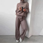 Femme en ensemble pull et pantalon en laine beige, tenant une tasse, appuyée contre un mur blanc. Mode décontractée, tenue confortable automne-hiver.