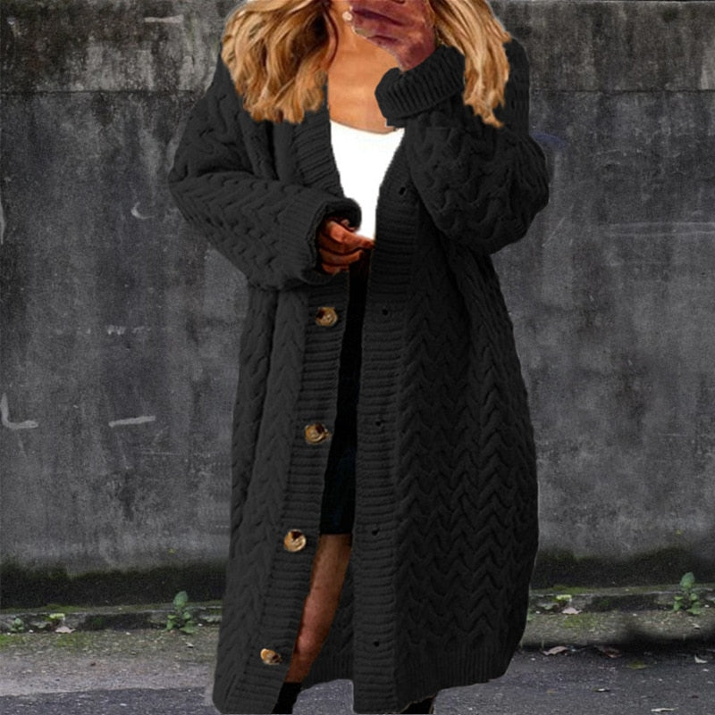 Femme portant un long cardigan noir à boutons dorés, sur fond de mur gris. Mode automne, style décontracté, tendance vêtements chauds.