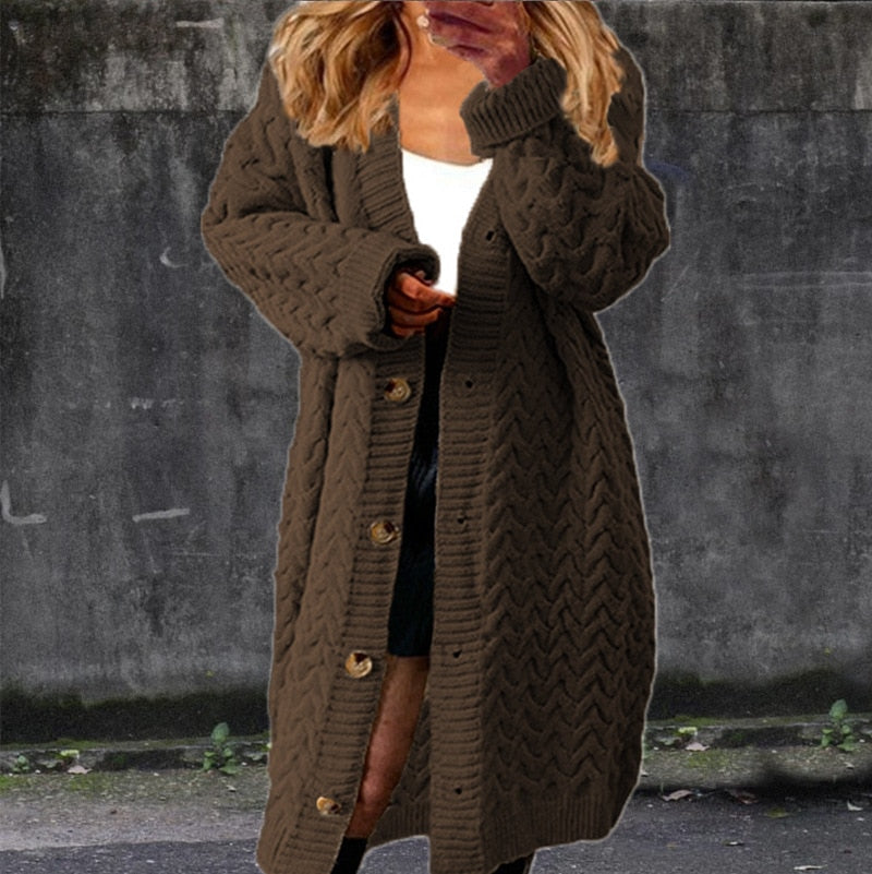 Femme portant un long cardigan marron à boutons, motif torsadé, sur un top blanc et jupe noire, posant devant un mur gris. Mode automne-hiver tendance.