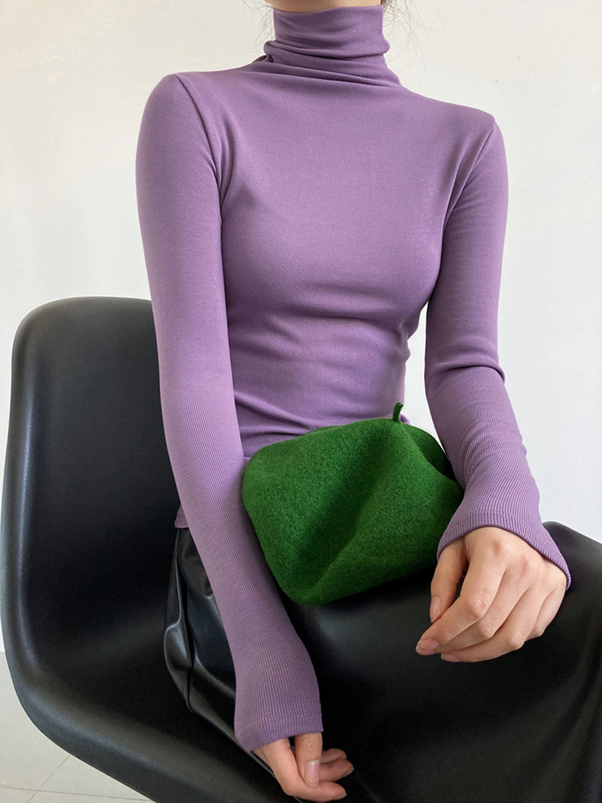 Femme en pull violet à col roulé, assise sur une chaise noire, tenant un sac vert. Mode automne, style élégant et moderne.