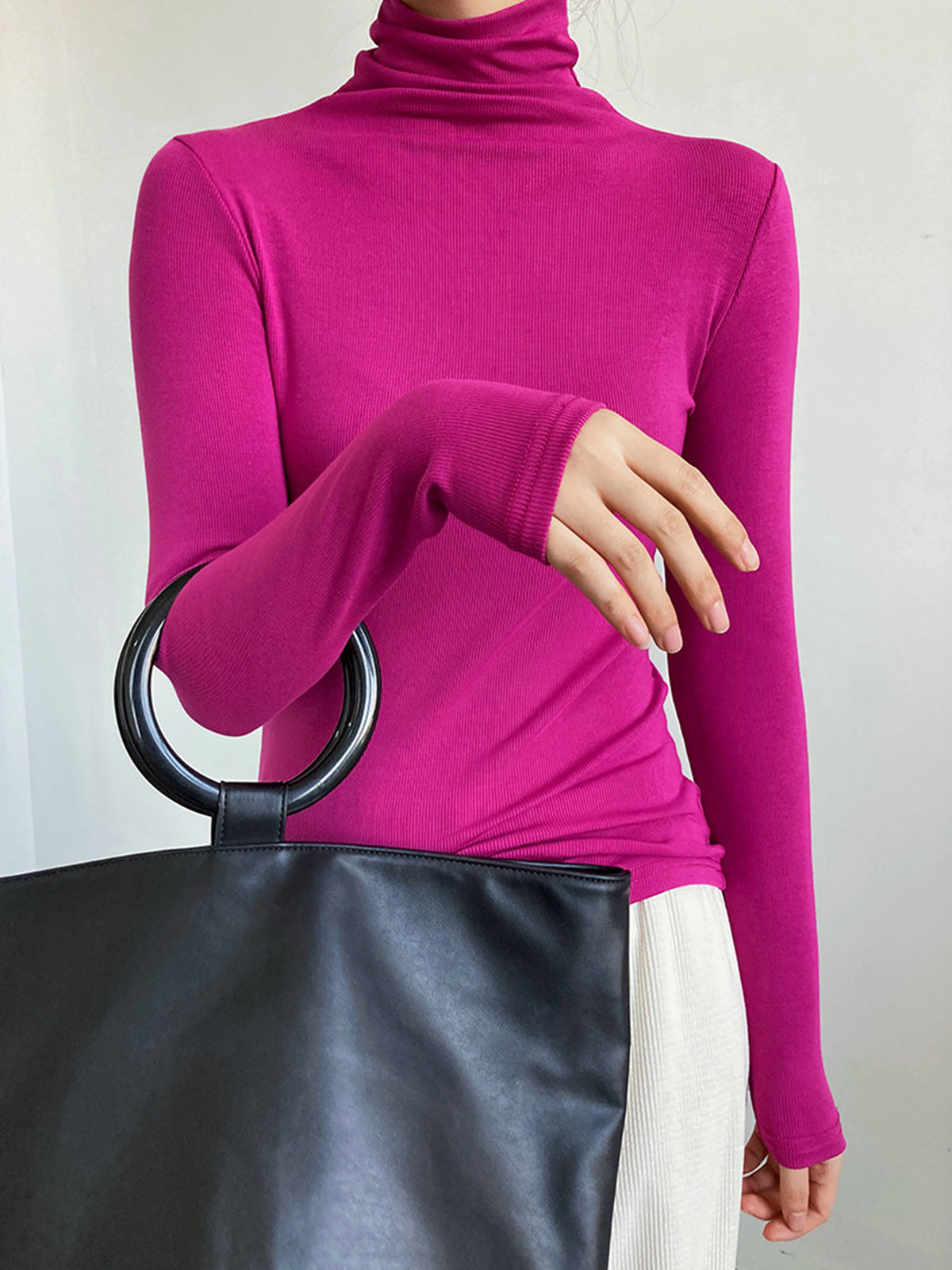 Femme portant un pull col roulé fuchsia, tenant un sac noir à anses rondes. Mode élégante, tenue automne-hiver, style moderne et chic.