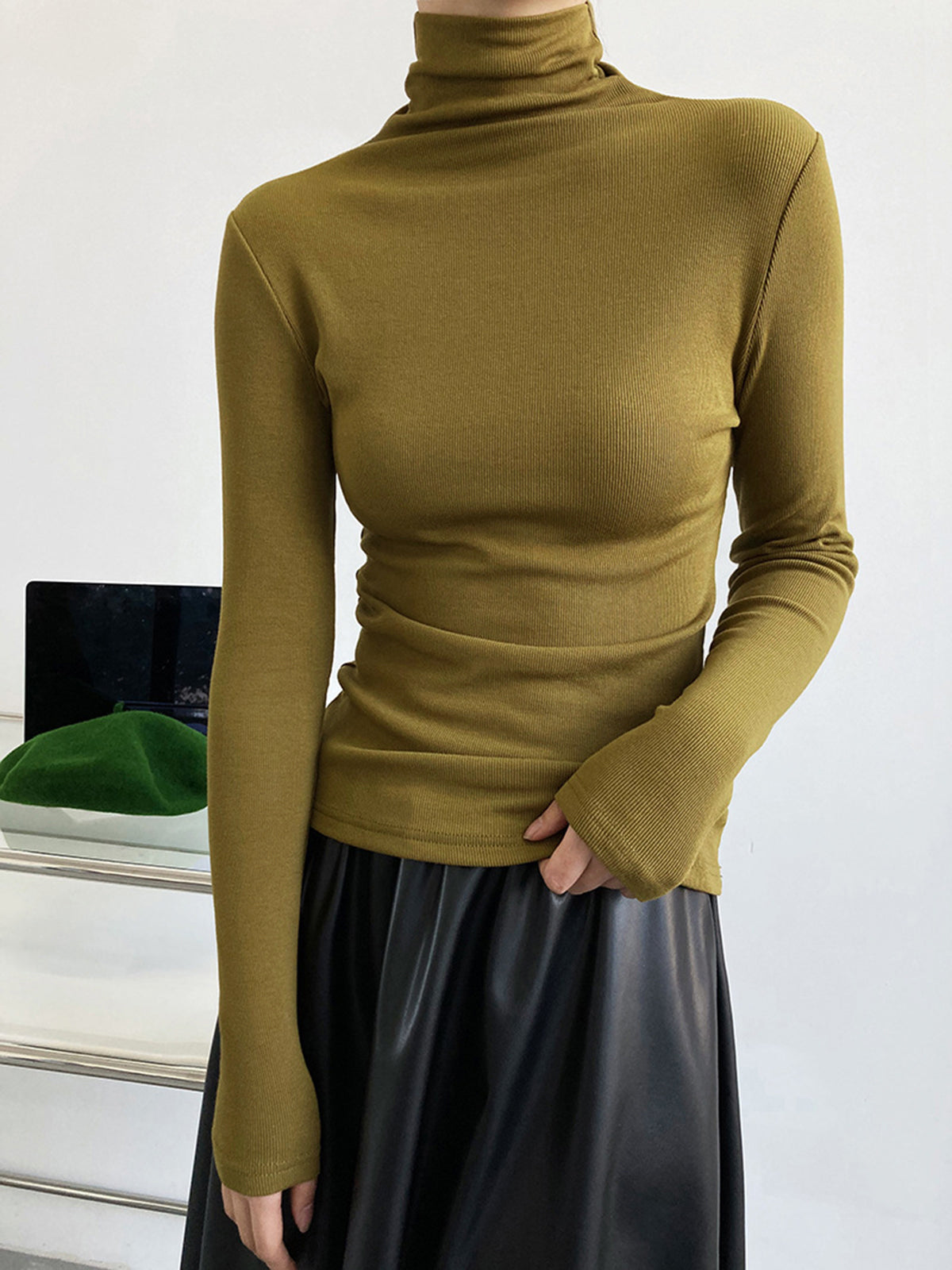 Femme portant un pull col roulé vert olive ajusté et une jupe noire en cuir, posant devant un fond minimaliste. Mode automne-hiver élégante.