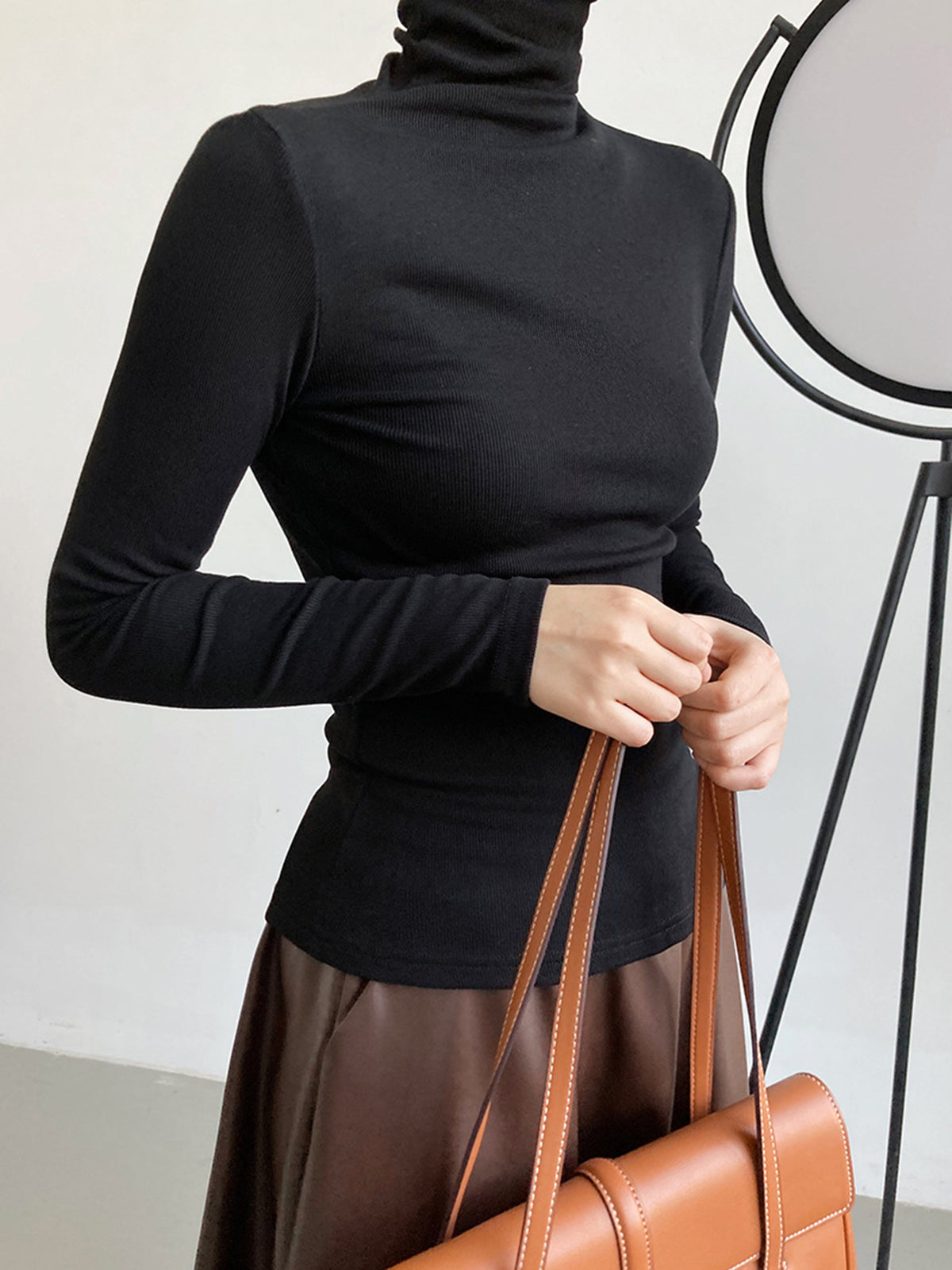 Femme portant un pull col roulé noir ajusté, tenant un sac à main marron. Mode élégante, tenue automne-hiver, style minimaliste.