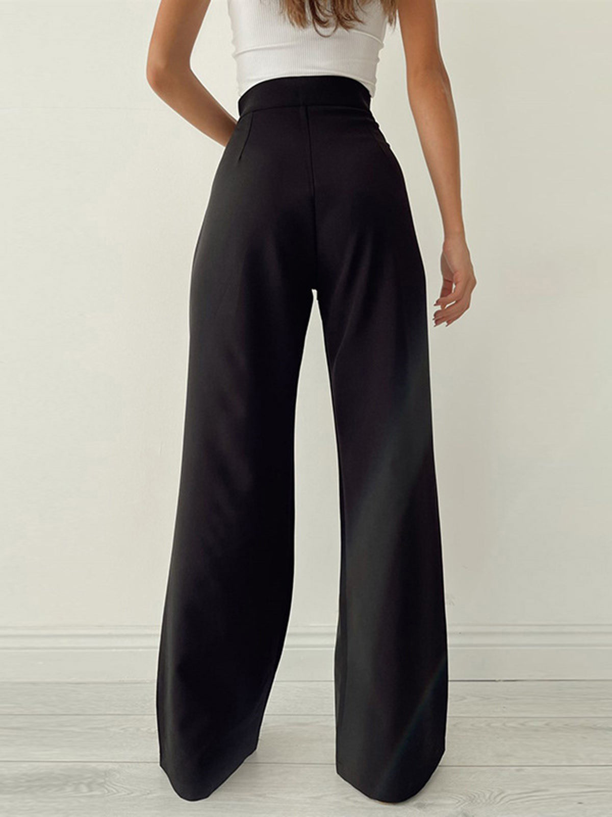 Femme portant un pantalon large noir taille haute, vue de dos, avec un haut blanc sans manches. Mode élégante et moderne, style minimaliste.