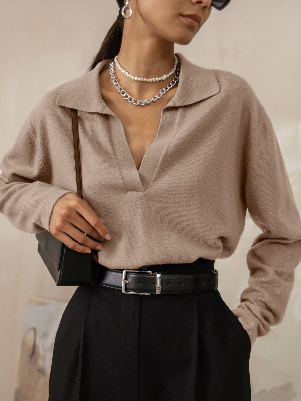 Femme portant un pull beige à col ouvert, collier en chaîne argentée, pantalon noir taille haute et sac à main noir. Mode élégante et moderne.