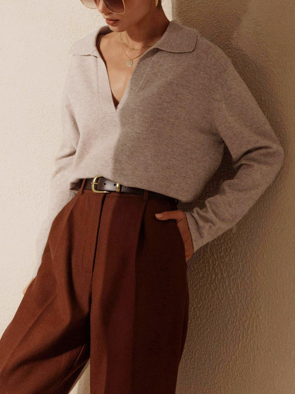 Femme portant un pull beige à col ouvert et un pantalon marron taille haute, accessoirisé d'une ceinture noire, posant contre un mur texturé.