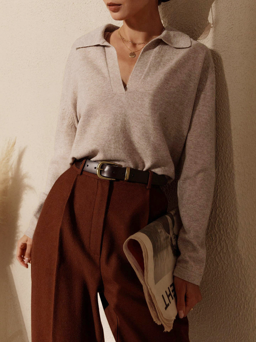Femme élégante portant un pull beige à col ouvert et un pantalon marron, tenant un journal. Mode automne chic, style décontracté sophistiqué.