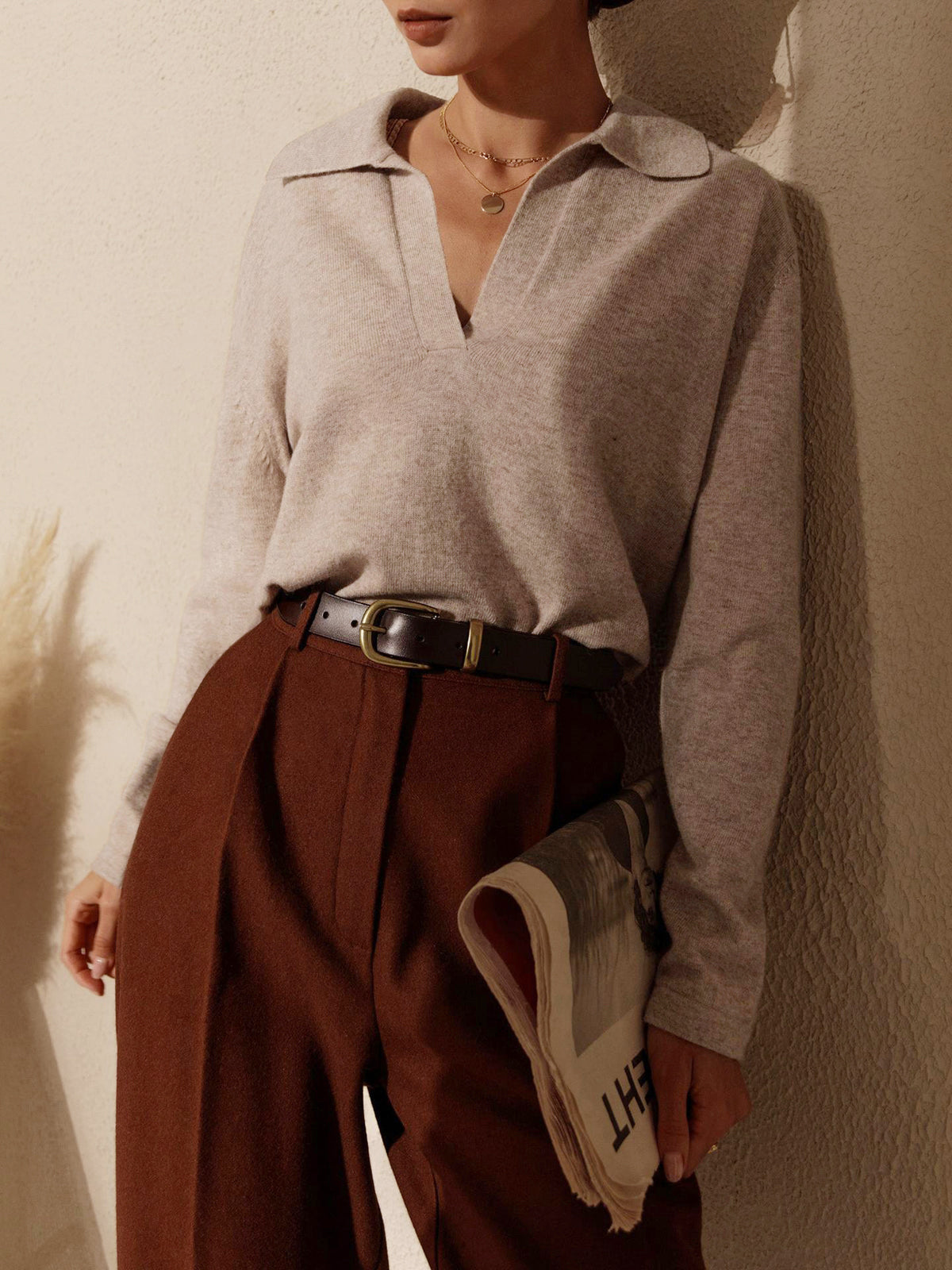 Femme élégante portant un pull beige à col ouvert et un pantalon marron, tenant un journal. Mode automne chic, style décontracté sophistiqué.