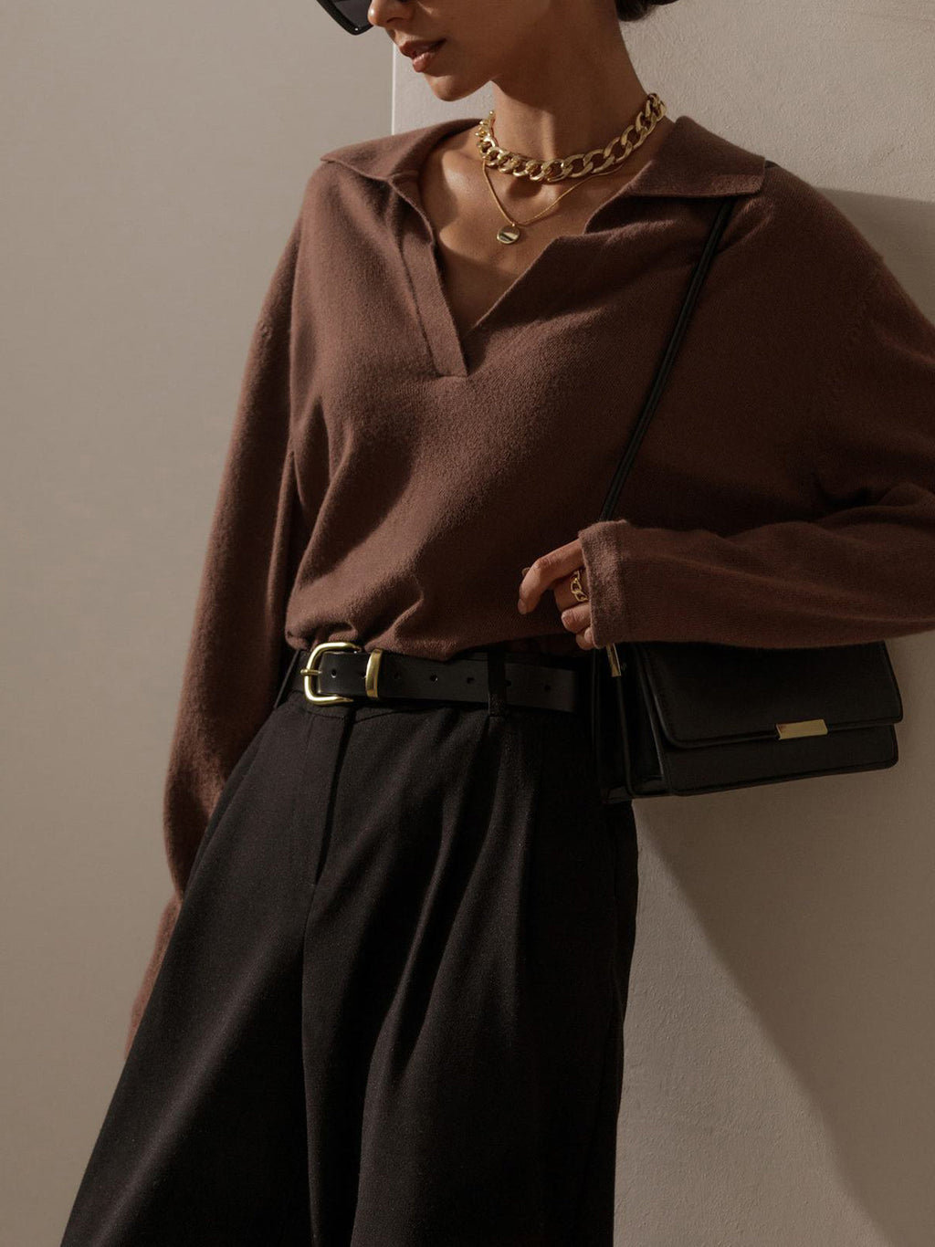 Femme élégante portant un pull marron, pantalon noir, ceinture en cuir, collier doré et sac à main noir. Mode chic et minimaliste.
