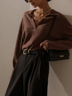 Femme élégante portant un pull marron, pantalon noir, ceinture en cuir, collier doré et sac à main noir. Mode chic et minimaliste.