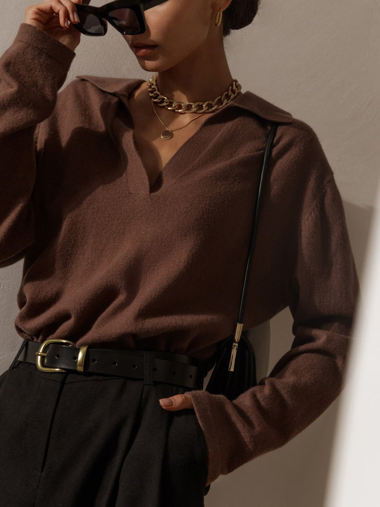 Femme élégante portant un pull marron, lunettes de soleil noires, collier doré, ceinture noire et pantalon sombre, posant contre un mur blanc.