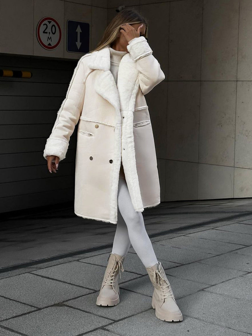 Femme portant un manteau en laine blanc, leggings blancs et bottes beiges, posant devant un mur moderne. Mode hiver élégante et tendance.