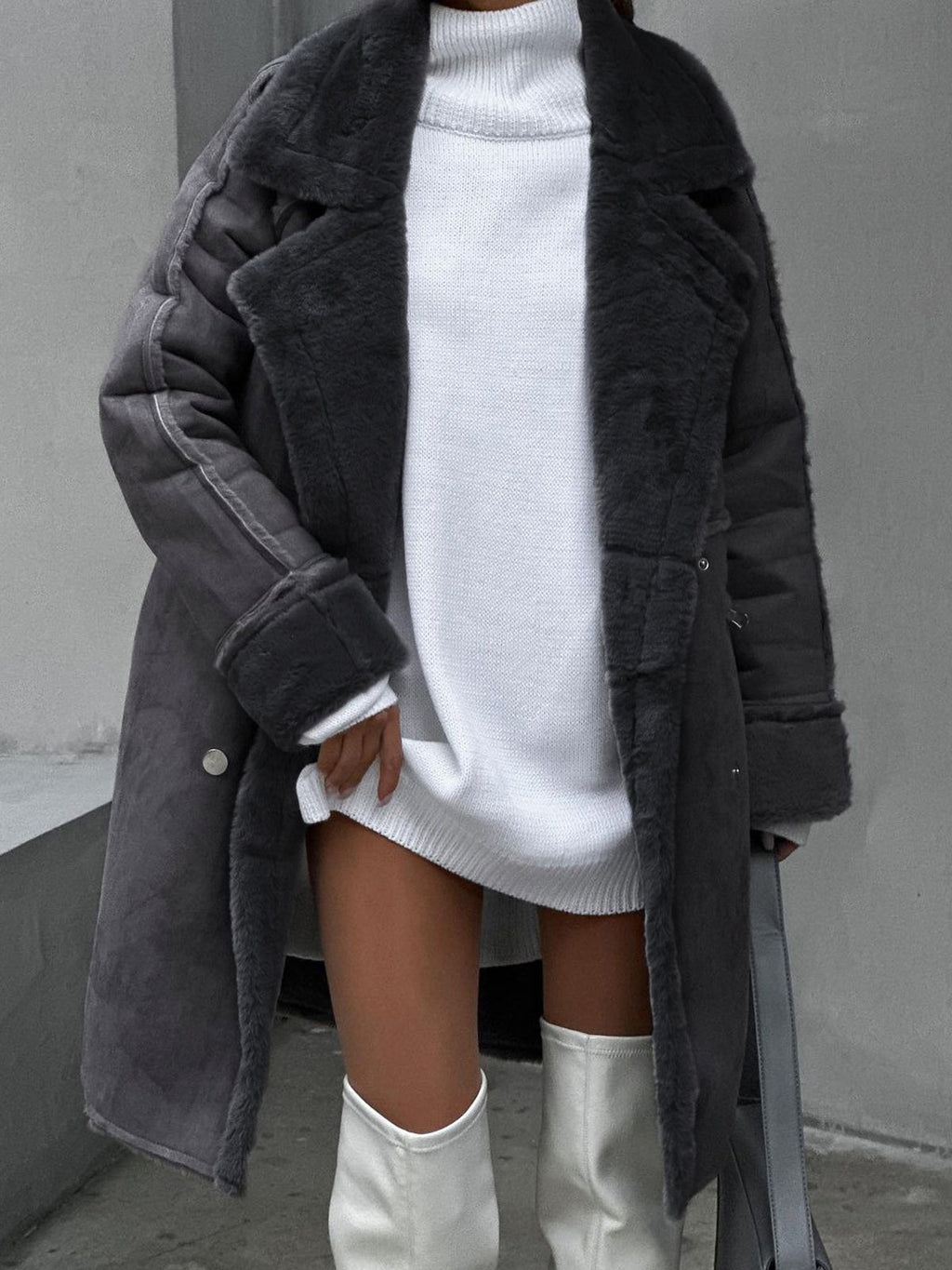 Femme portant un manteau en fausse fourrure gris sur une robe pull blanche, avec des bottes hautes blanches. Mode hiver élégante et tendance.
