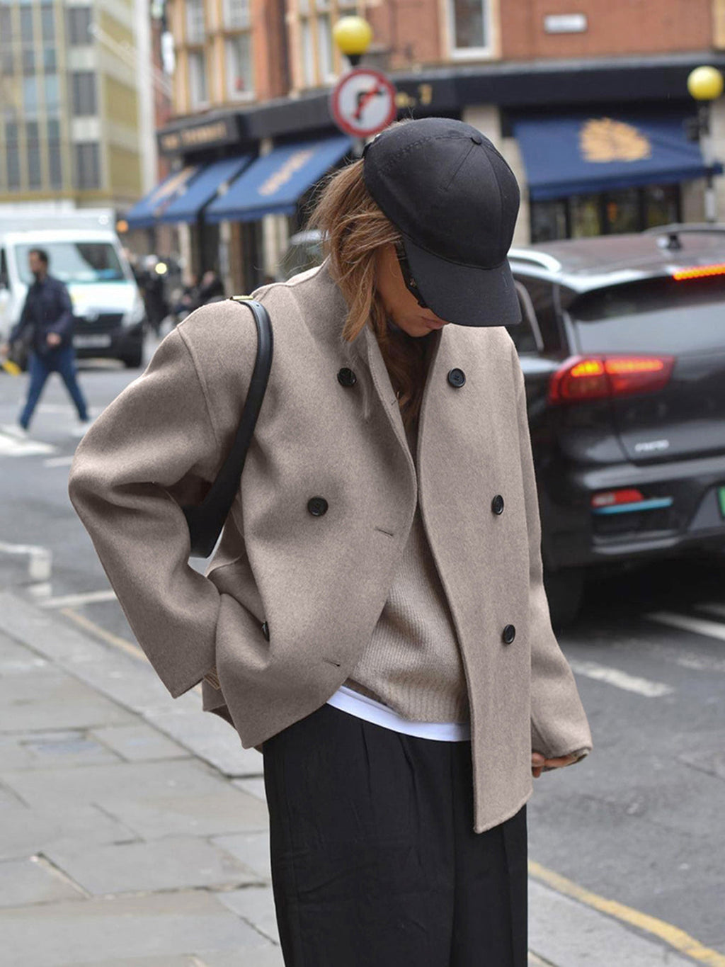 Femme portant un manteau beige, casquette noire, et sac à bandoulière, marchant en ville. Mode urbaine, style décontracté, automne.