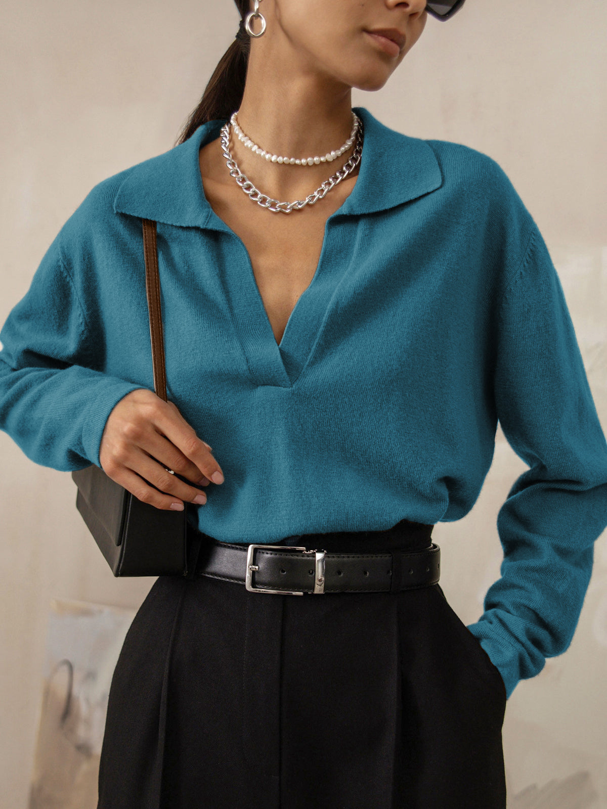 Femme portant un pull bleu col ouvert, collier chaîne argentée, pantalon noir taille haute, sac à main noir. Mode élégante et moderne.