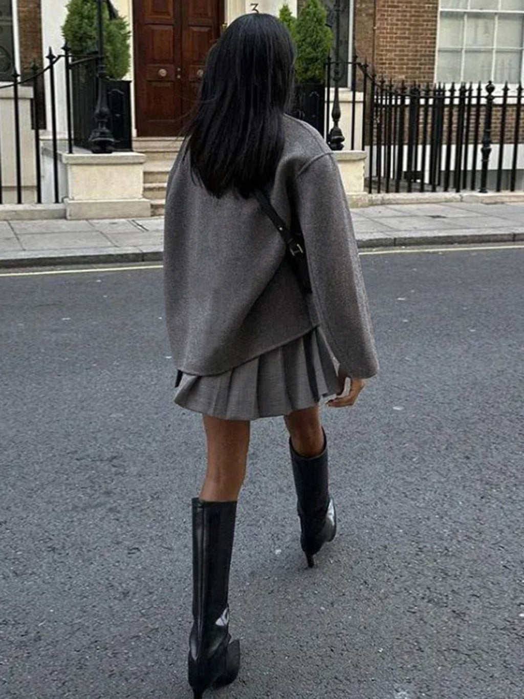 Femme en jupe plissée grise, pull oversize et bottes noires marchant dans une rue urbaine. Mode automne, style urbain chic, tenue décontractée.