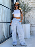 Femme en t-shirt blanc et pantalon rayé bleu, posant devant un mur en pierre avec lunettes de soleil, style décontracté chic, mode estivale.