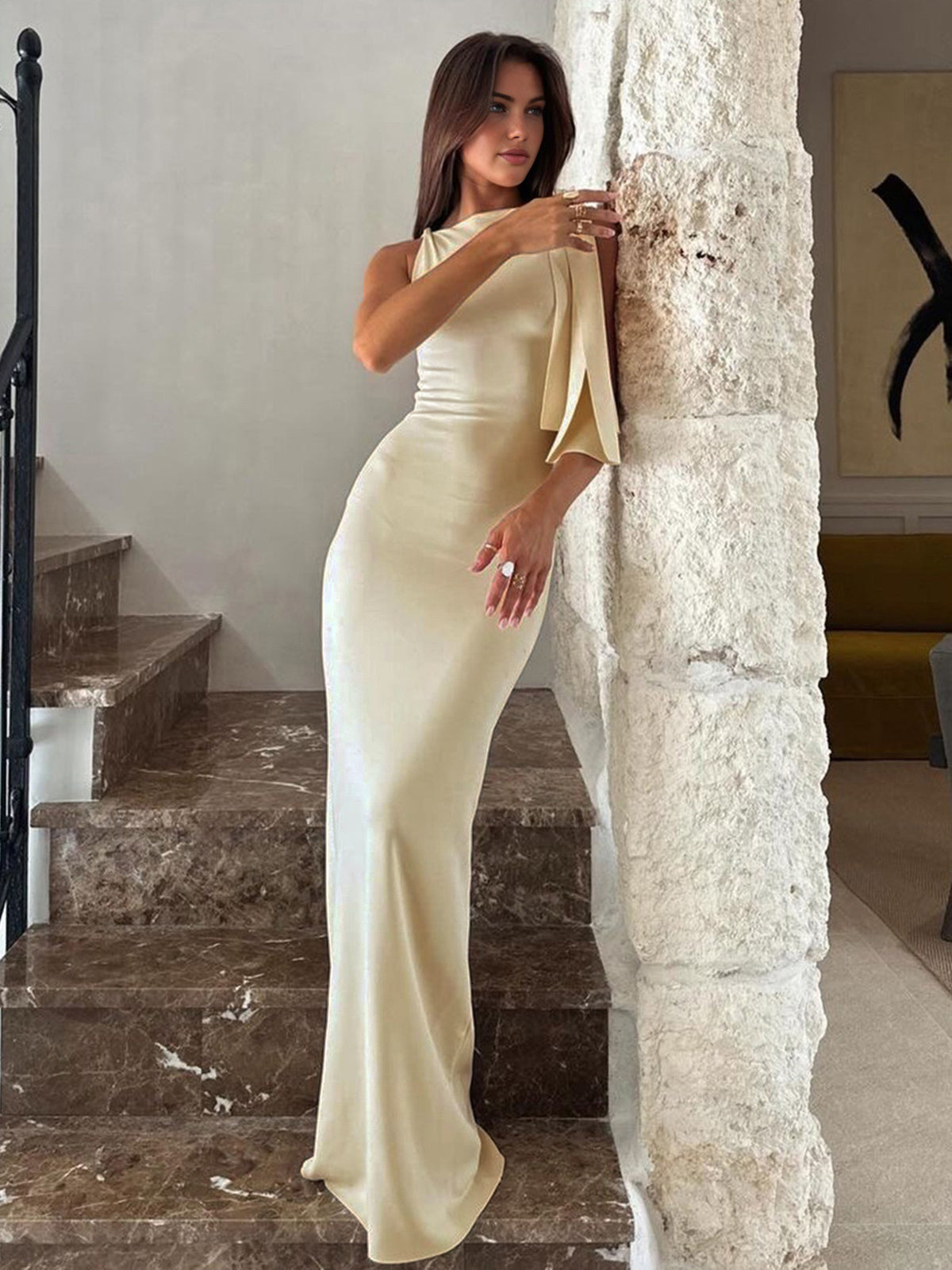 Femme élégante en robe longue beige satinée, posant près d'un mur en pierre, intérieur moderne. Mode féminine, style chic, robe de soirée.
