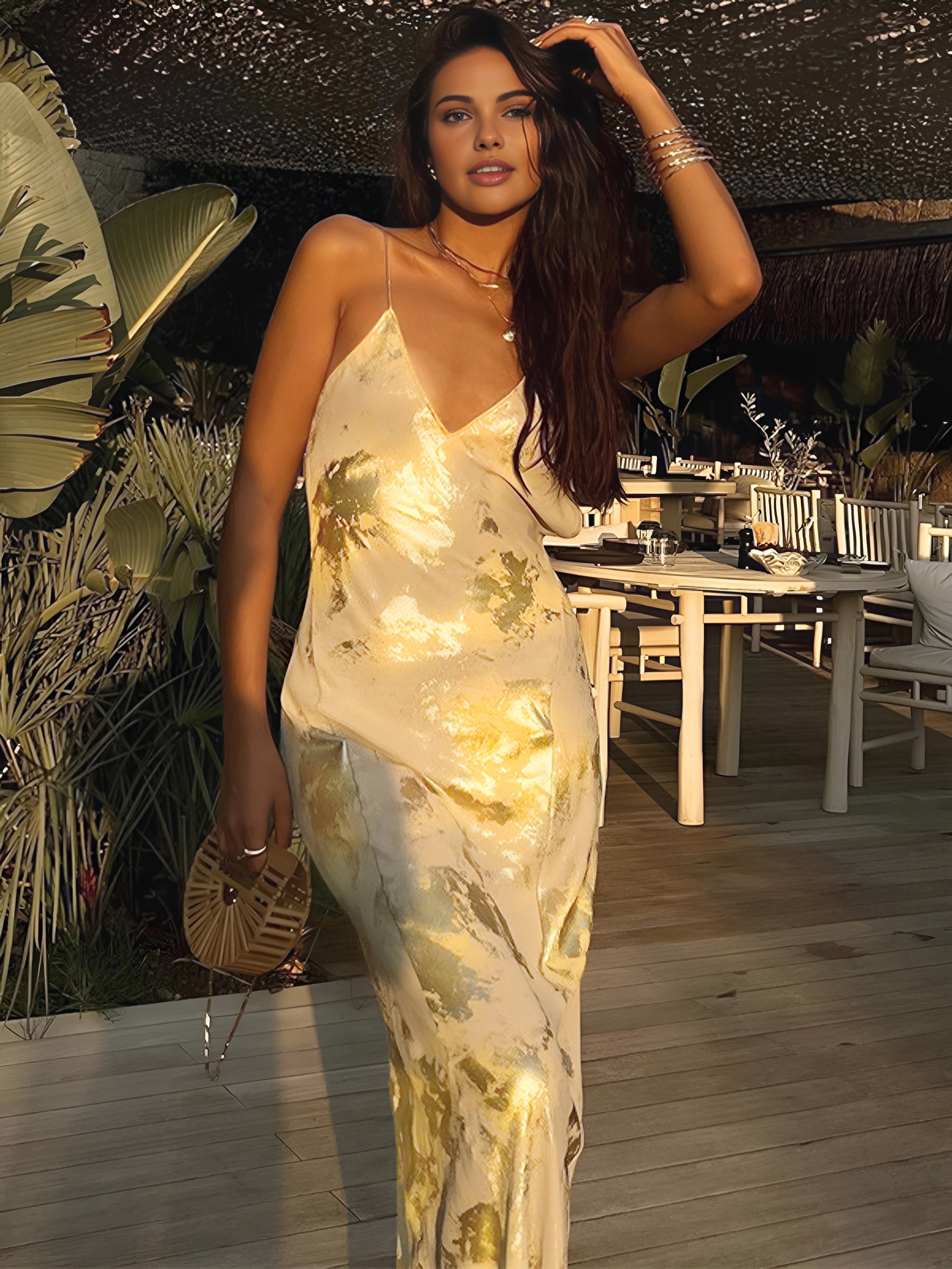 Femme élégante en robe dorée satinée, posant sur une terrasse en bois avec plantes exotiques et mobilier de jardin, ambiance coucher de soleil.