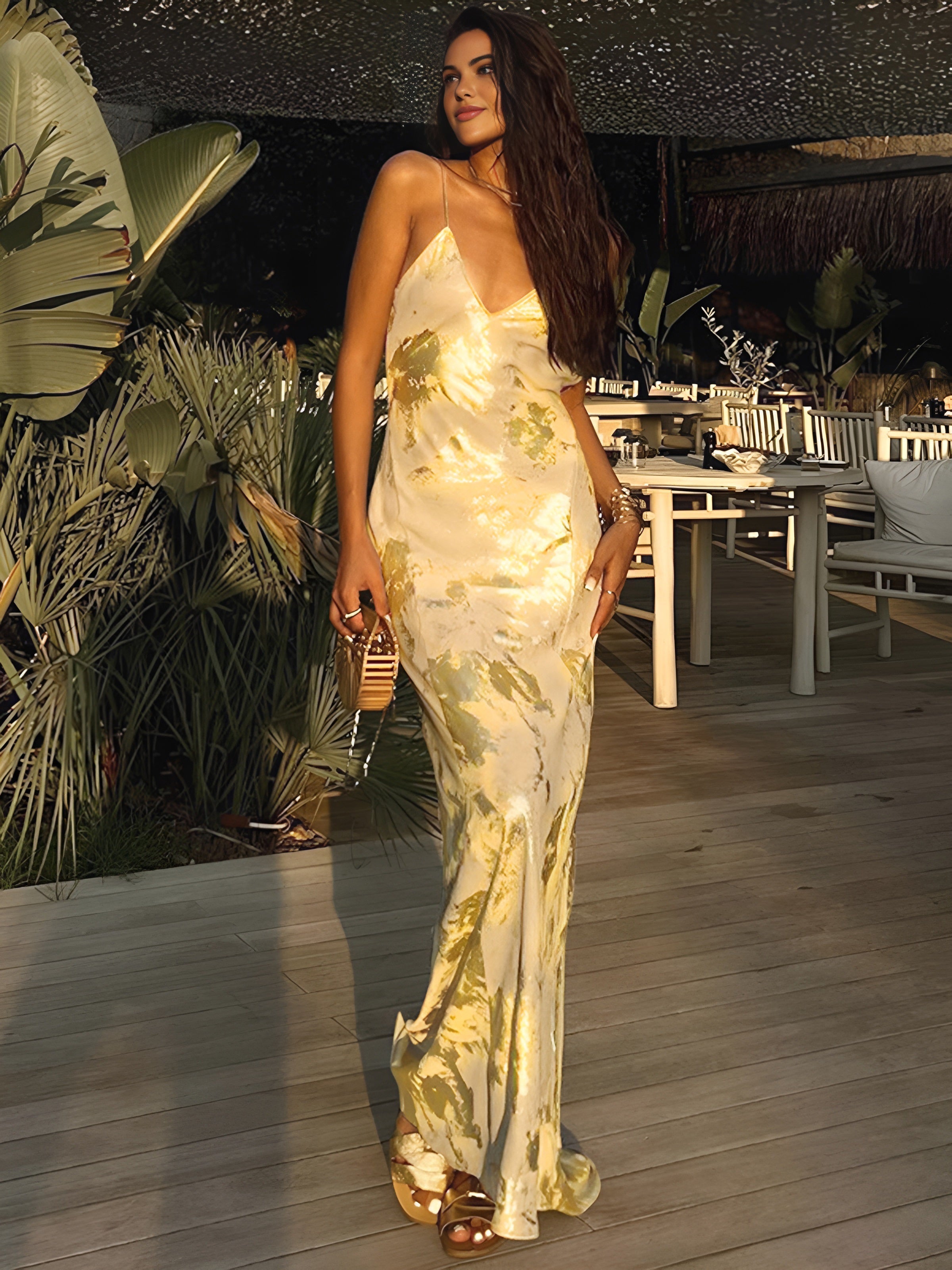 Femme élégante en robe longue satinée à motifs floraux, posant sur une terrasse en bois, entourée de plantes exotiques, ambiance soirée chic.