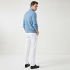 Homme portant un pull bleu en laine torsadée et un pantalon blanc, debout de dos dans un studio minimaliste. Mode masculine élégante.