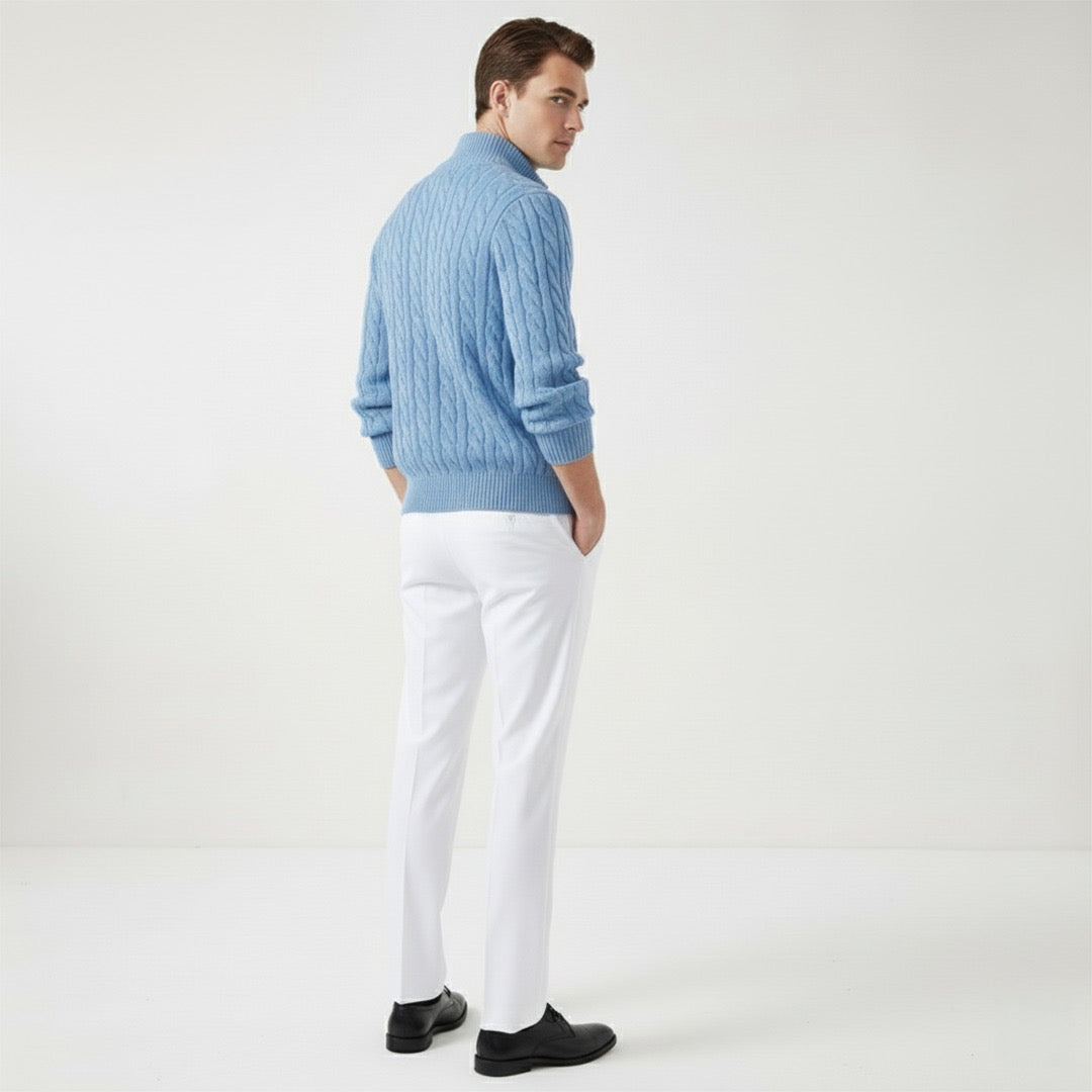 Homme portant un pull bleu en laine torsadée et un pantalon blanc, debout de dos dans un studio minimaliste. Mode masculine élégante.