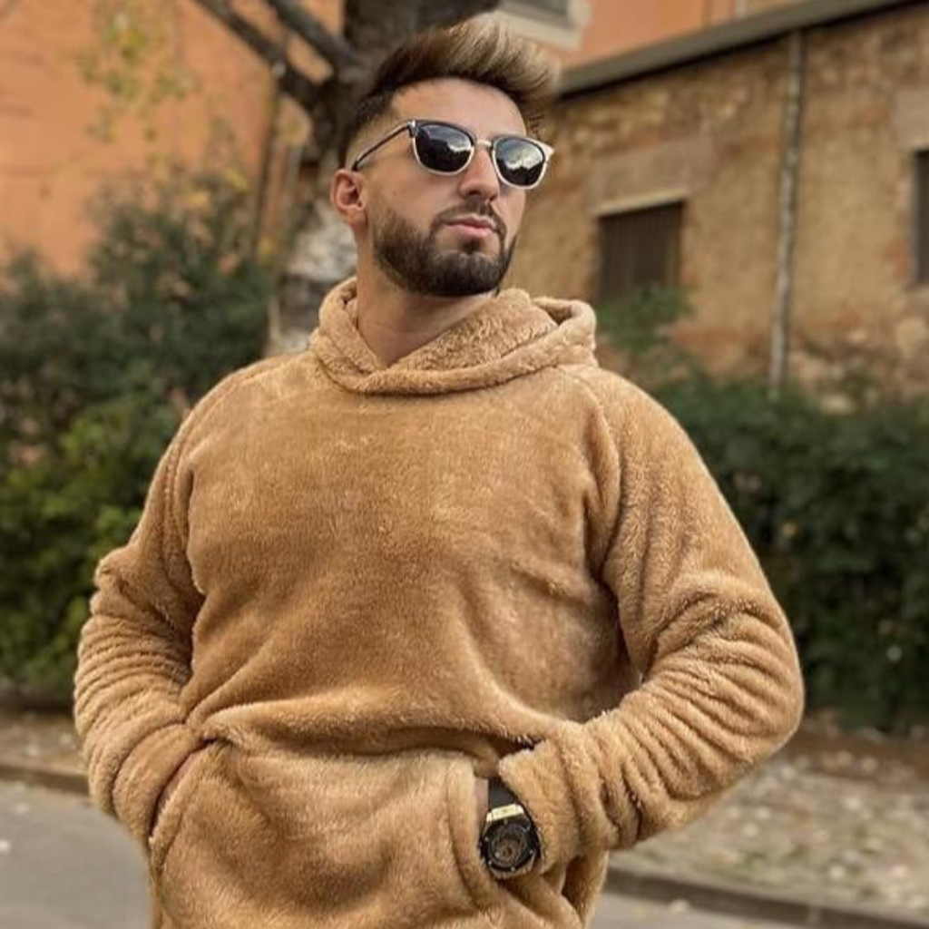 Homme barbu portant un sweat à capuche en peluche beige et des lunettes de soleil, posant à l'extérieur devant un bâtiment en brique.