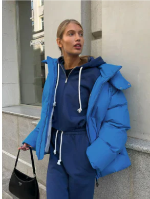 Femme en doudoune bleue et survêtement bleu, tenant un sac à main noir, debout devant un bâtiment urbain. Mode hiver, style décontracté.