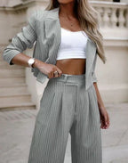 Femme en tailleur pantalon rayé gris, top blanc, montre élégante, posant devant un bâtiment classique. Mode chic, tendance, style urbain.