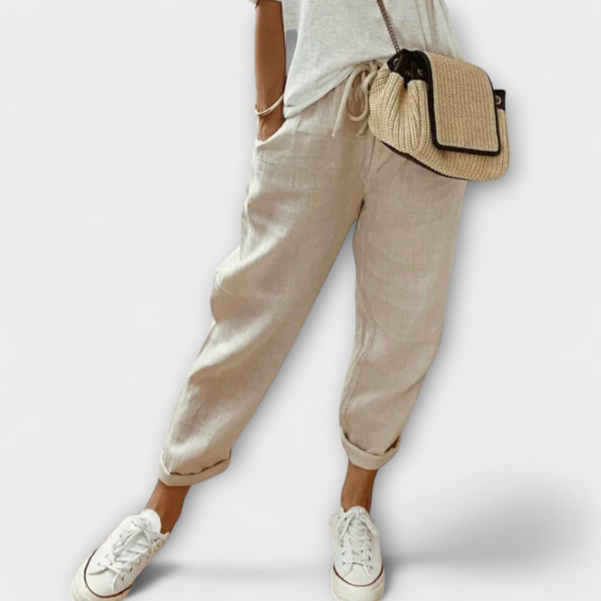 Pantalon en lin beige pour femme, style décontracté, avec poches, associé à un t-shirt blanc et des baskets blanches, sac en osier en bandoulière.