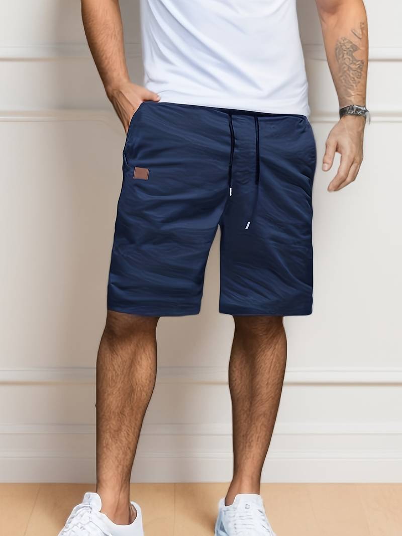 Homme portant un short bleu marine décontracté avec cordon de serrage, t-shirt blanc et baskets blanches, posant devant un mur intérieur.