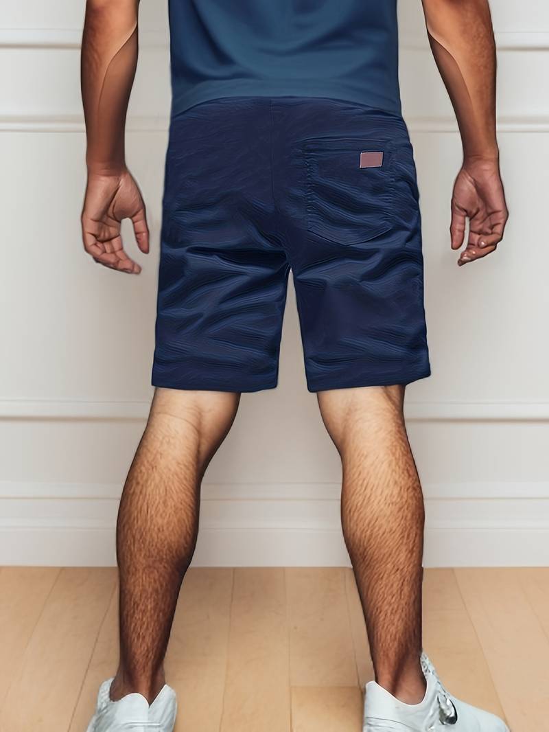 Homme en short bleu marine et t-shirt bleu, debout de dos, sur un sol en bois clair, portant des baskets blanches. Mode décontractée masculine.