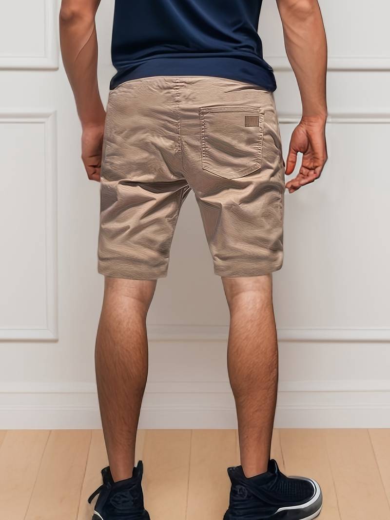 Homme en short beige et baskets noires, debout de dos devant un mur blanc. Mode masculine décontractée, tenue estivale, style moderne.