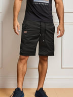 Homme portant un short noir décontracté et un t-shirt noir, debout sur un sol en bois clair. Mode masculine, tenue estivale, style décontracté.
