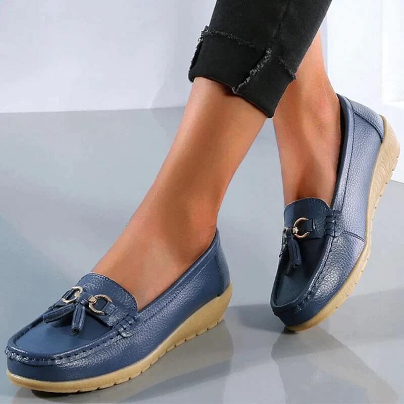 Chaussures mocassins bleues pour femme avec semelles compensées beiges, ornées de pampilles et boucles métalliques, portées avec un pantalon noir.
