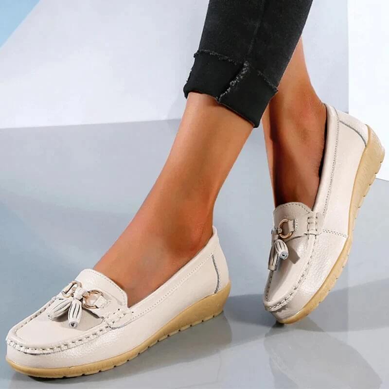 Mocassins blancs pour femme avec pompons, semelle beige antidérapante, portés avec un pantalon noir. Chaussures élégantes et confortables.