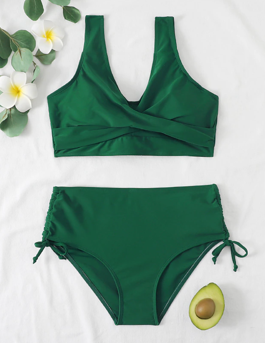 Maillot de bain vert deux pièces avec haut croisé et culotte taille haute, décoré de fleurs blanches et demi-avocat sur fond blanc.